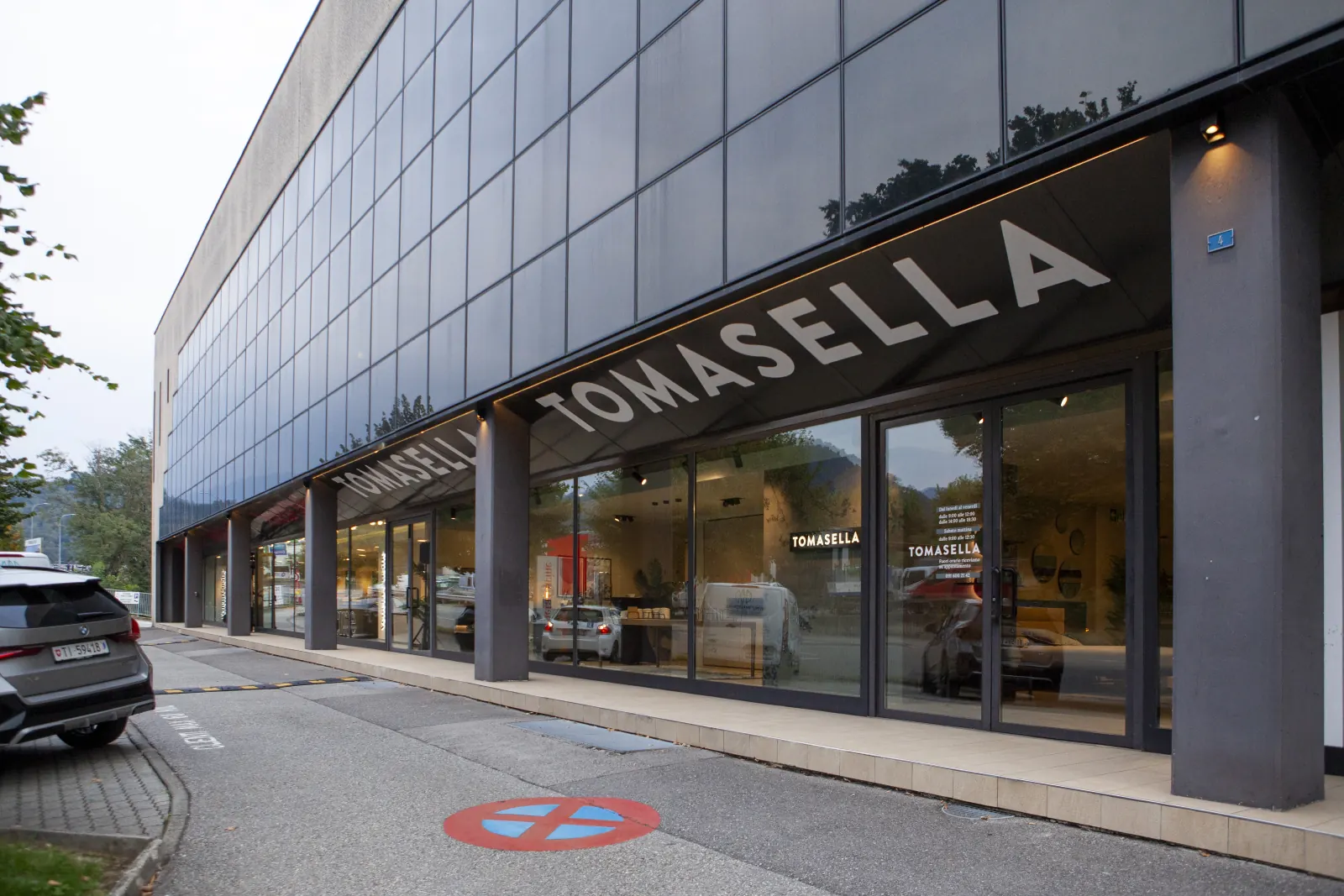 Ingresso dello Store Tomasella di Manno visto dalla strada, con l’insegna TOMASELLA sulla pensilina e le vetrine illuminate